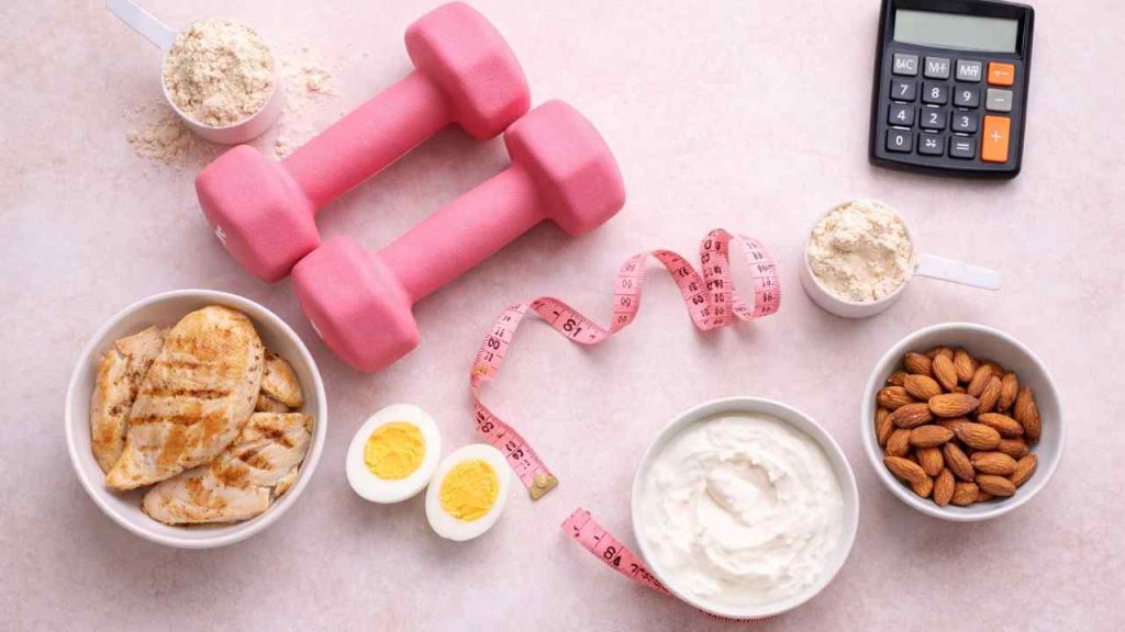 Flat lay of high-protein foods including chicken breast, boiled eggs, Greek yogurt, almonds, whey protein powder, pink dumbbells, measuring tape, and calculator on soft pink background.
